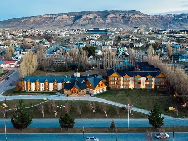 Colorful wooden architecture overlooks a blue river in a Patagonian town nestled beneath dramatic plateaus.