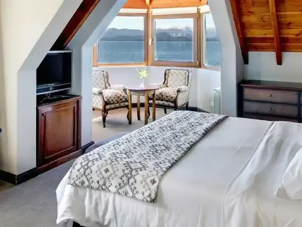 Luxury hotel bedroom in Bariloche with lake views, featuring a queen bed, sitting area with armchairs, and panoramic windows overlooking Patagonian landscape.