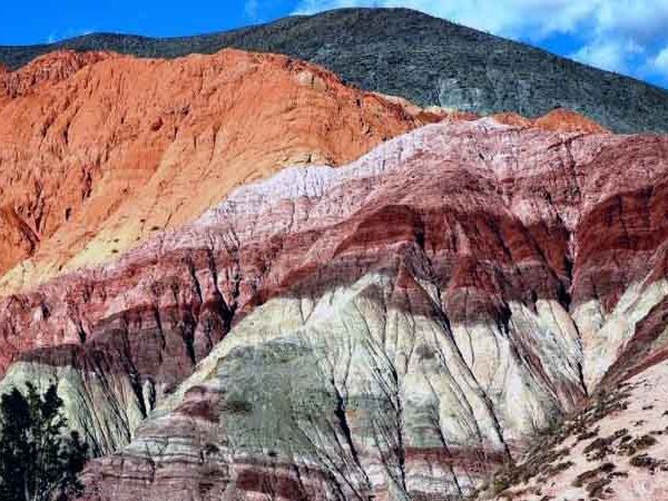 Colorful striped mountain hillside displays vibrant layers of red, orange, white, and green rock formations in Salta, Argentina.