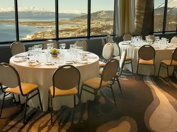 Elegant banquet hall at Arakur Ushuaia with round tables overlooking the Beagle Channel and snow-capped mountains.