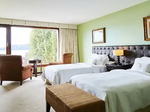 Luxury hotel bedroom with twin beds, leather headboard, and panoramic views of Patagonian lakes and mountains through floor-to-ceiling windows.