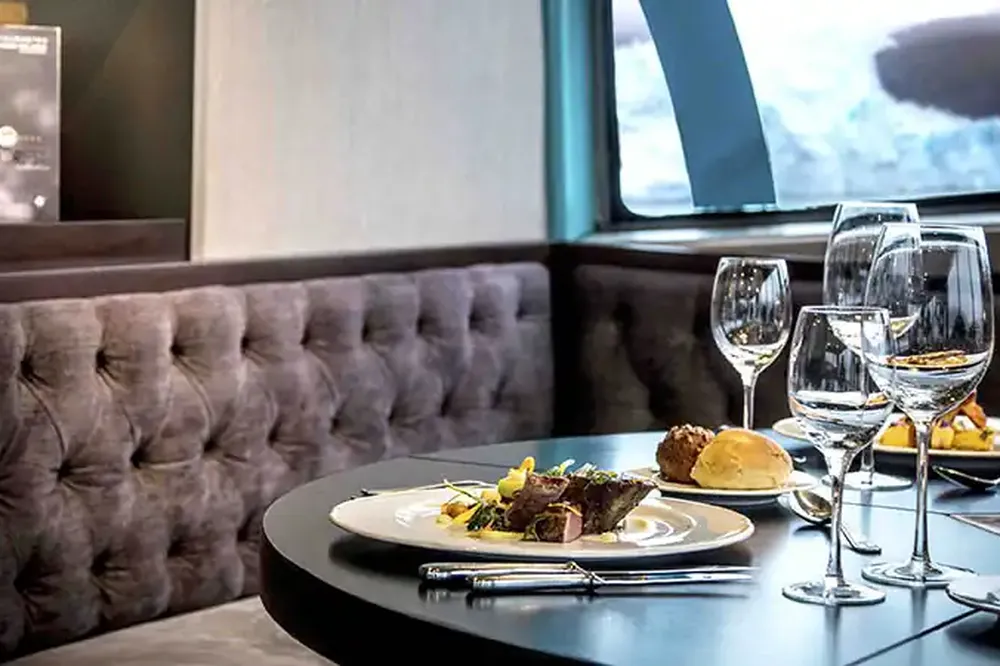 Elegant fine dining setup overlooking Patagonian glaciers with plated cuisine, wine glasses, and tufted seating.
