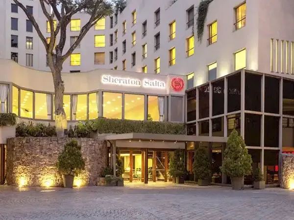 Sheraton Salta hotel entrance with illuminated courtyard, stone facade, and ivy-covered residential tower at dusk.