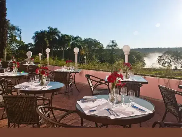 Elegant outdoor dining terrace overlooking Iguazú Falls with red flowers, white linens, and lush subtropical forest.