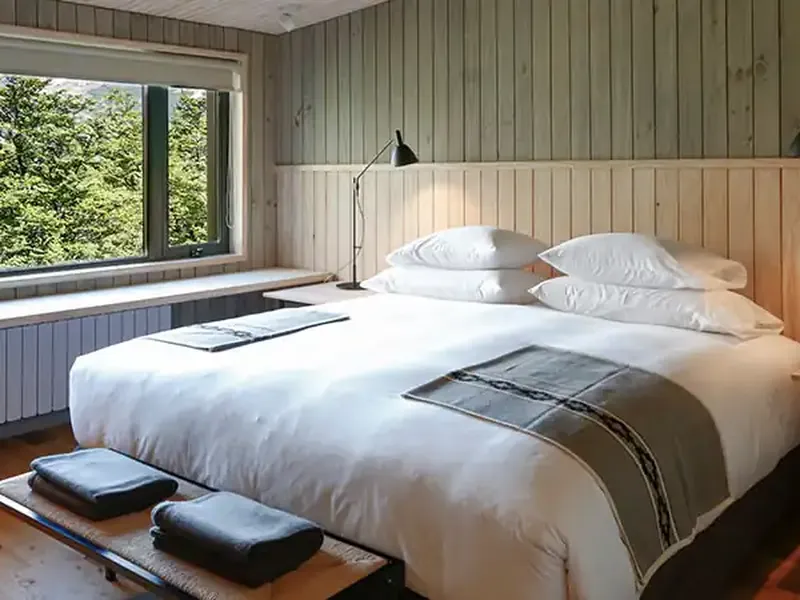 Modern lodge bedroom with mountain views, white bedding, wood paneling, and large windows overlooking Patagonian landscape.