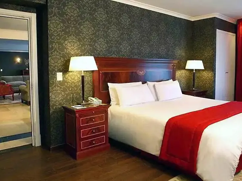 Luxurious hotel bedroom suite with damask wallpaper, mahogany furnishings, and red velvet accents in Buenos Aires.