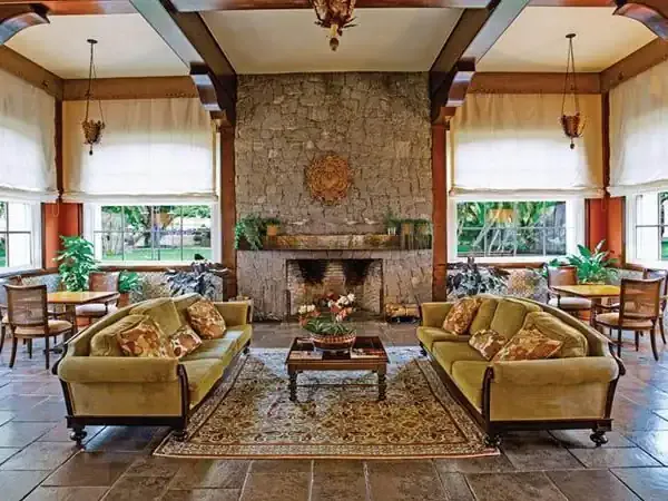 Elegant hotel lounge with symmetrical gold sofas, stone fireplace, and expansive windows overlooking gardens at Belmond luxury lodge.