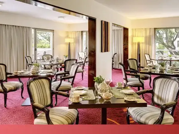 Elegant dining room at Sheraton hotel with striped upholstered chairs, multiple tables set for service, and pink carpeting overlooking gardens.