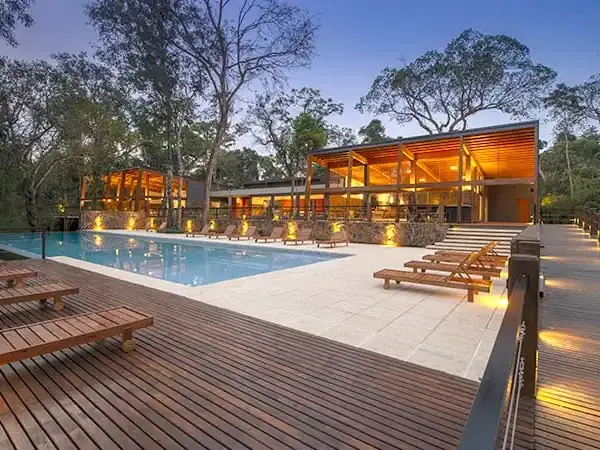 Luxury lodge with illuminated wooden architecture, infinity pool, and deck surrounded by native forest at dusk.