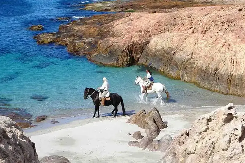Horseback riders wade through turquoise waters at a secluded Patagonian beach cove surrounded by dramatic rocky cliffs.