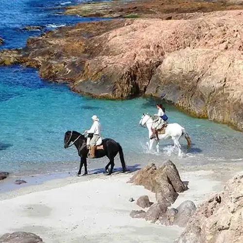 Horseback riders wade through turquoise waters at a secluded Patagonian beach cove surrounded by dramatic rocky cliffs.