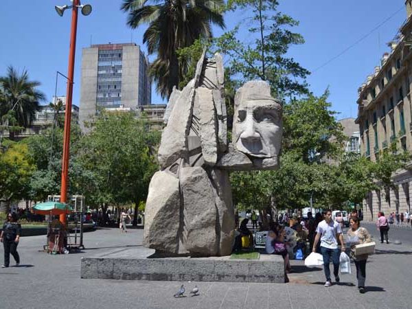 Large stone sculpture of a face in Plaza de Armas, Santiago de Chile's historic downtown square surrounded by colonial architecture.