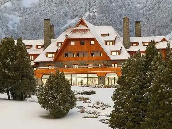 Llao Llao Hotel's distinctive wooden architecture and red roof covered in snow with snow-capped Andes mountains in the background.