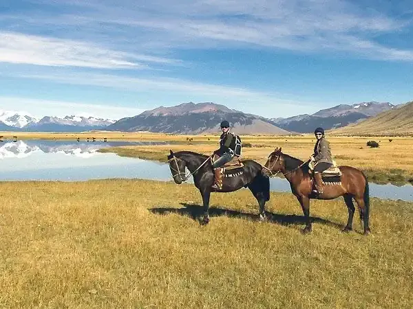 Two horseback riders explore the vast Patagonian steppe near Estancia Eolo, with snow-capped Andes mountains and pristine lagoons reflecting the sky.