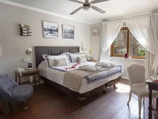 Elegant bedroom at Ecohotel Carmenere featuring a queen bed, dark wood furniture, white linens, and large windows overlooking Patagonian landscape.