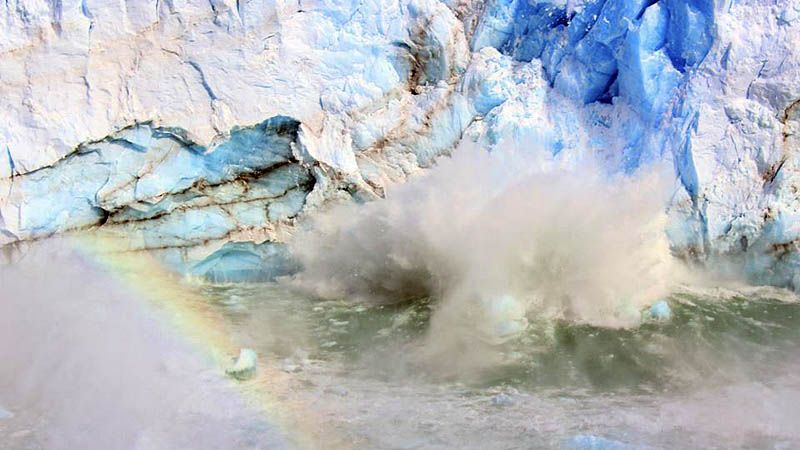 Massive blue glacier calving into water, creating a large splash with a faint rainbow visible in the mist. Perito Moreno Glacier