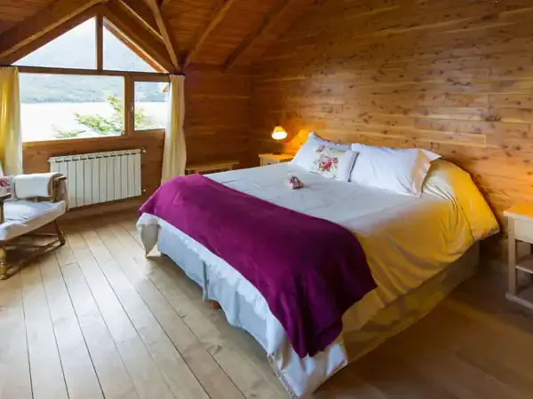 Rustic wooden lodge bedroom with purple and yellow bedding, large windows overlooking Patagonian landscape, and cozy furnishings.