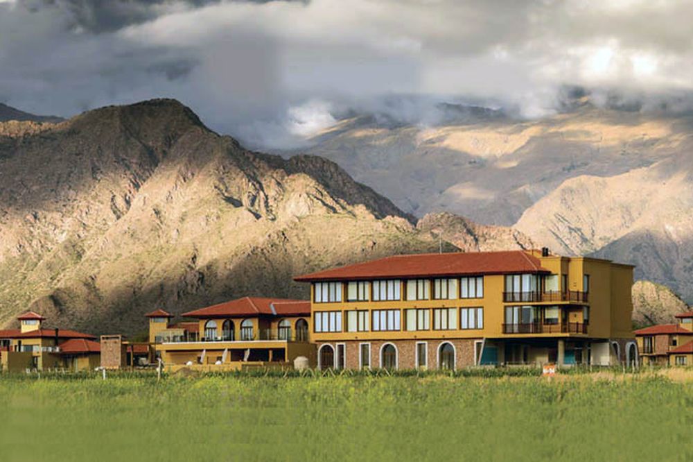 Colorful colonial-style lodge nestled in Cafayate's green valley with dramatic mountain backdrop and cloudy sky.