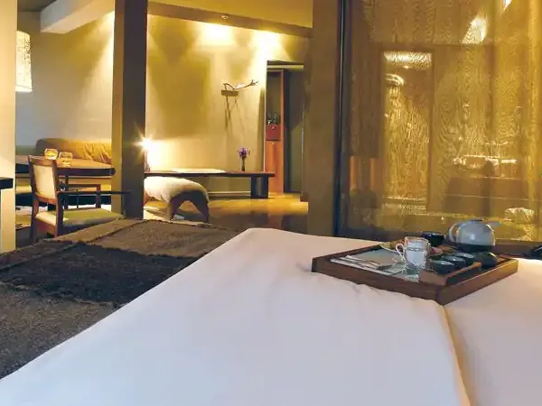 Luxurious modern hotel suite with warm lighting, white bed, dark furnishings, and panoramic windows overlooking a contemporary lounge area.