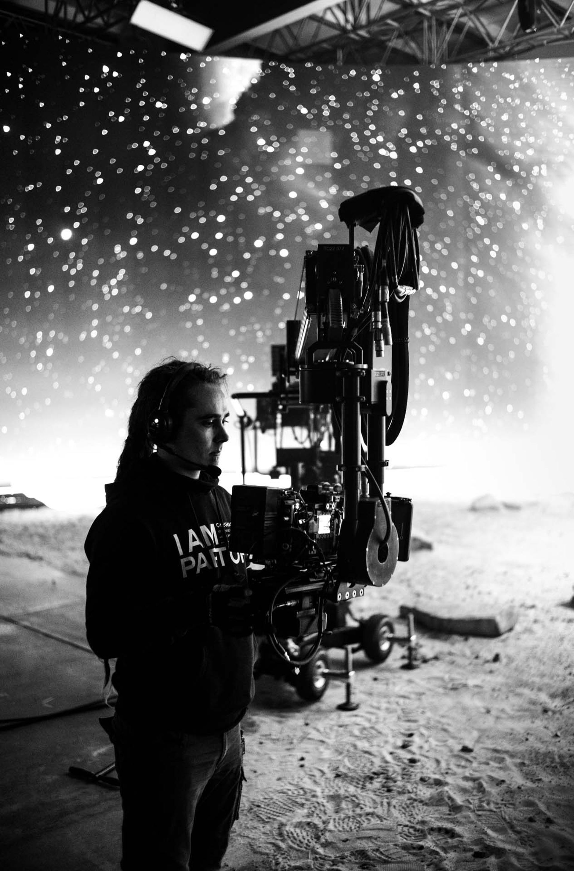 A person operates a large camera on a film set resembling a starry night sky, with sandy terrain and film equipment visible.