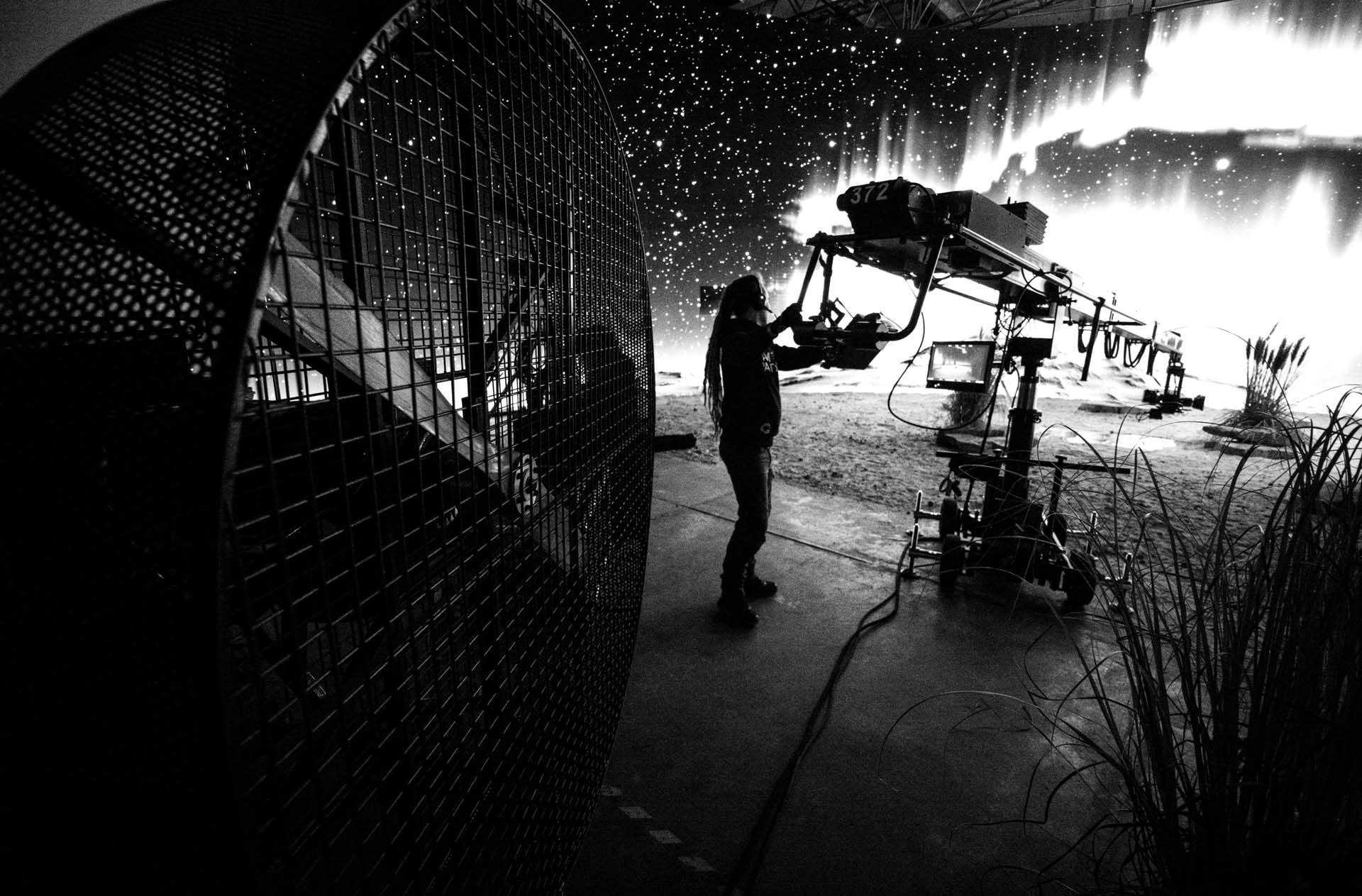 A person operates a large camera crane on a film set with floodlights in the background, creating a dramatic atmosphere.
