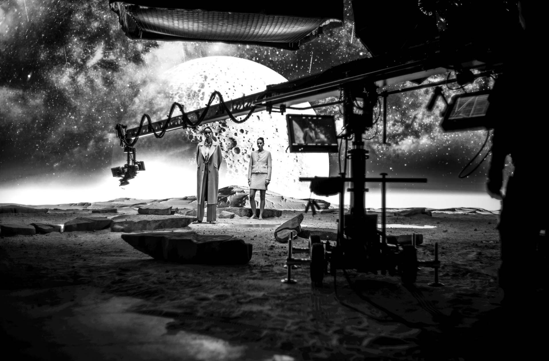 Black and white film set with two people, extensive camera equipment, and a cosmic backdrop featuring a large moon.