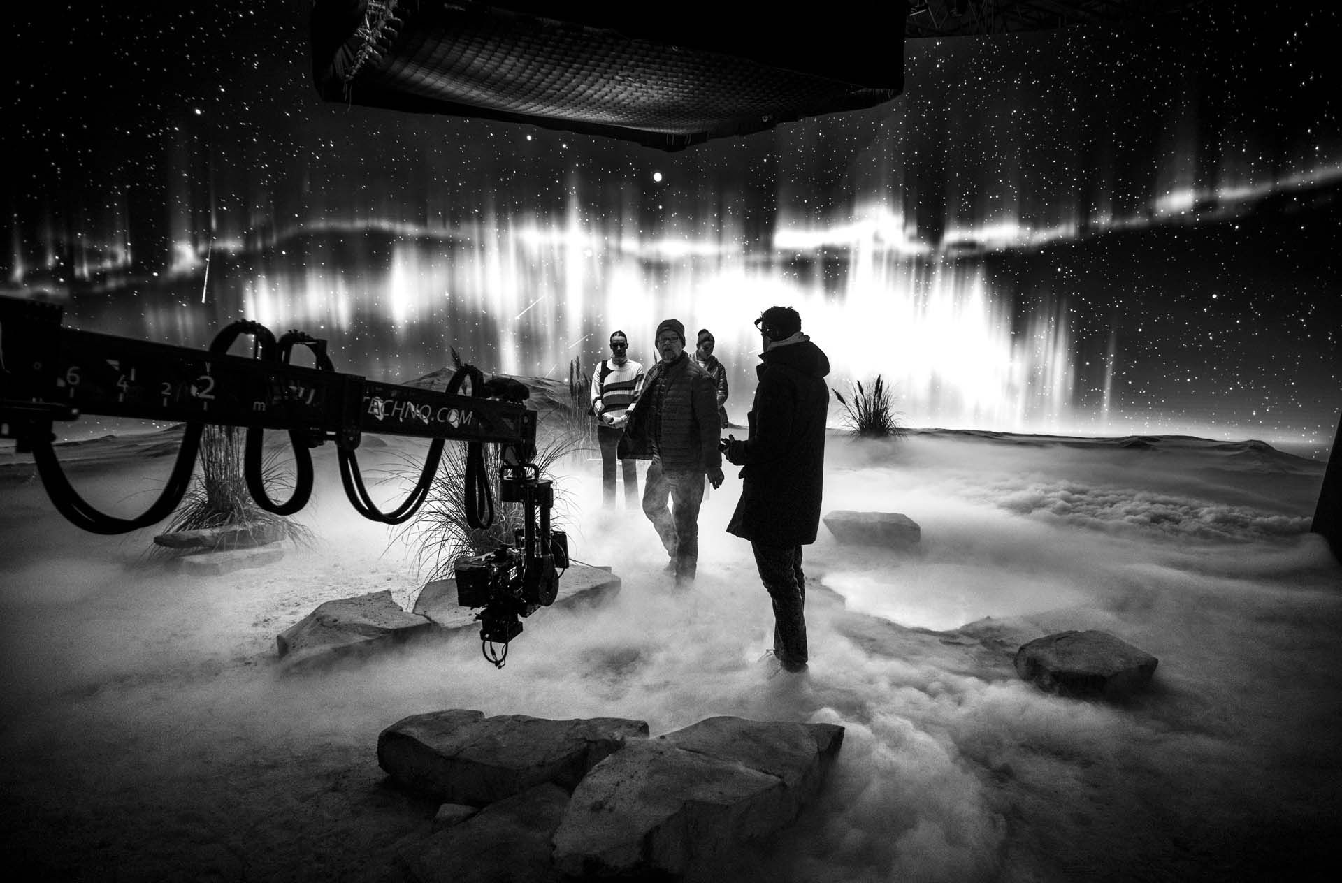 A film crew stands on a mist-covered set with rocks and a backdrop resembling a starry sky and aurora. A camera crane is visible on the left.