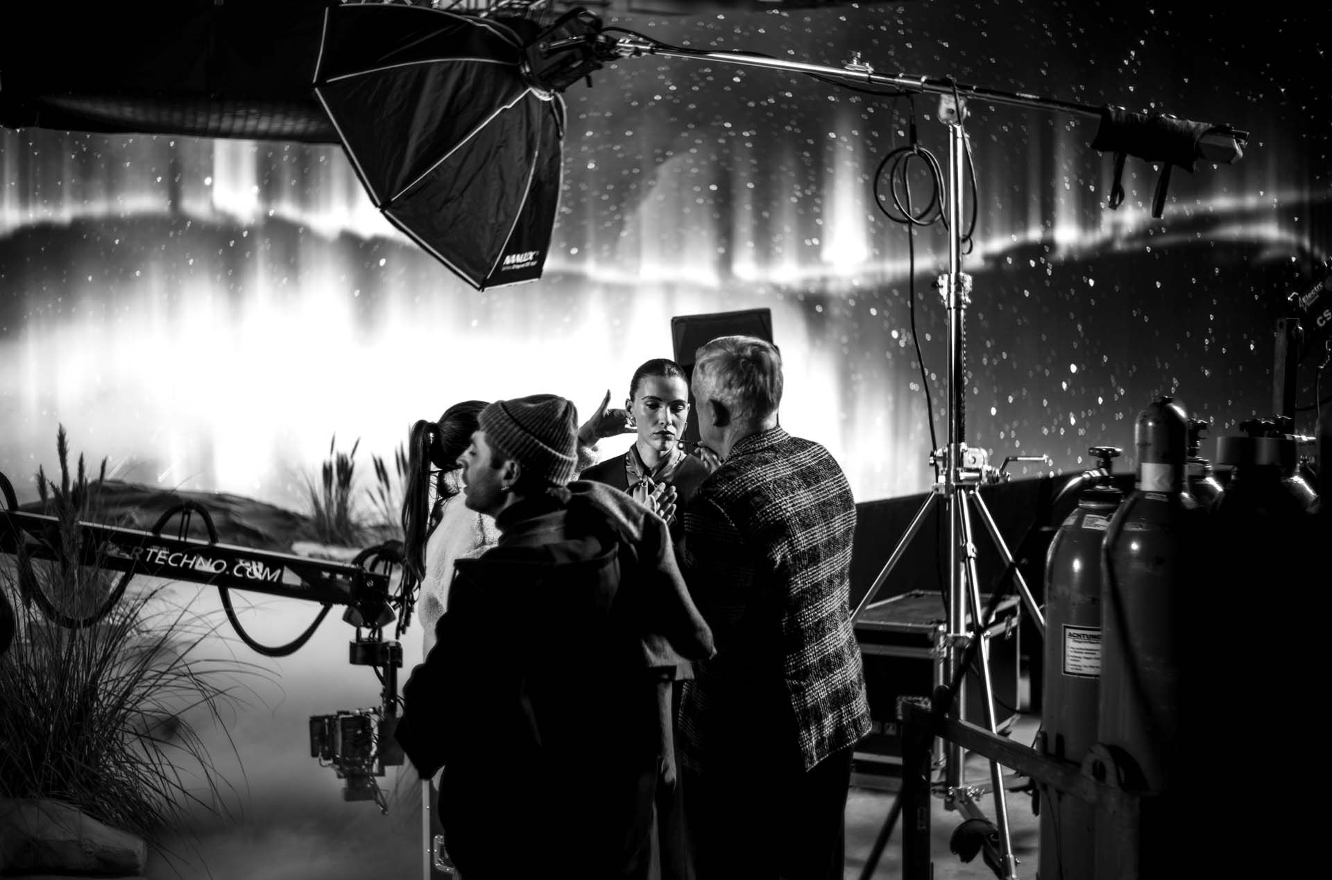 Black and white photo of a film crew on a set with lighting equipment and a northern lights backdrop, engaged in discussion.
