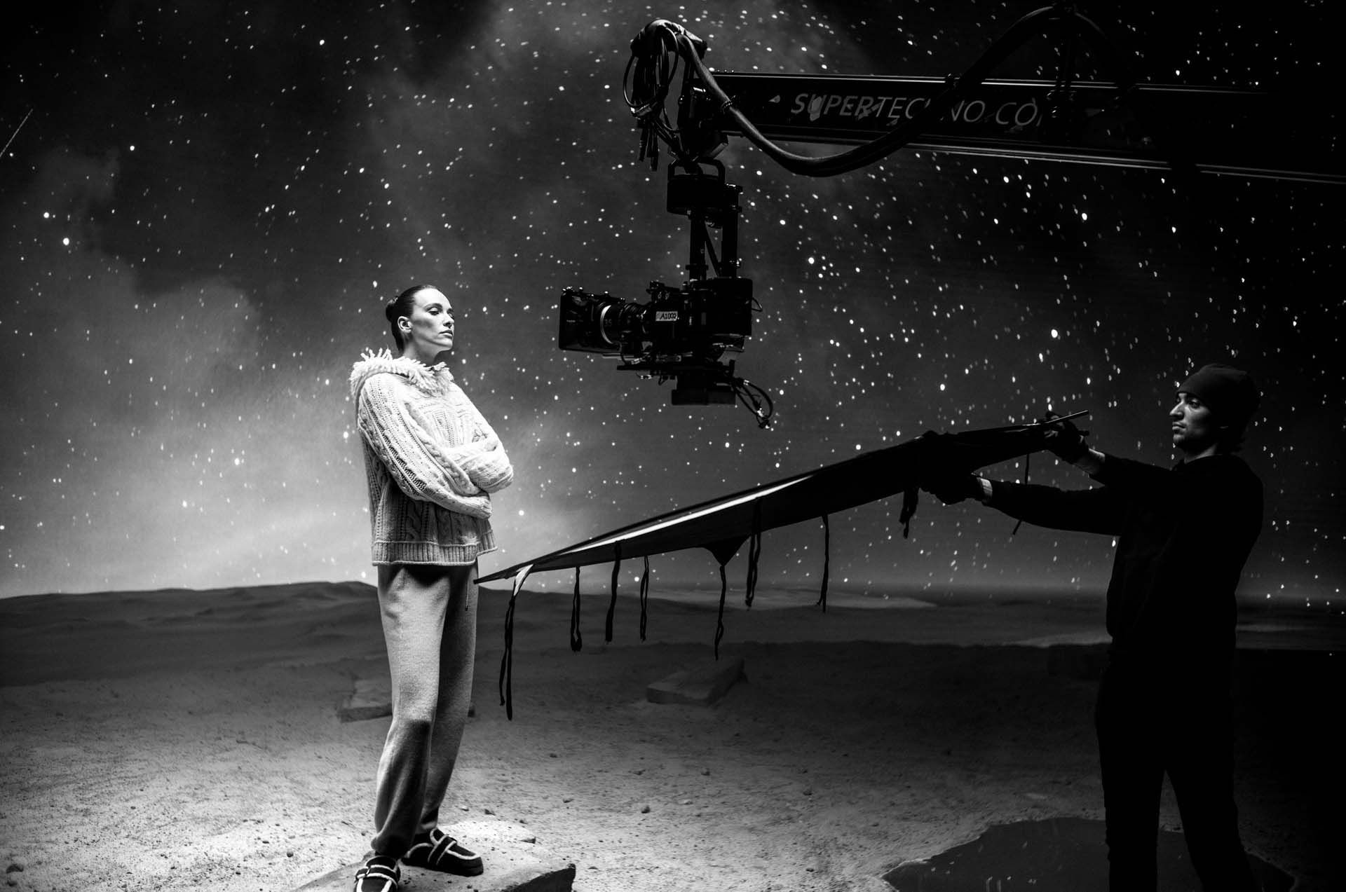 Black and white image of a person on a film set, with a camera crane and crew member holding equipment. Starry backdrop creates a night scene.
