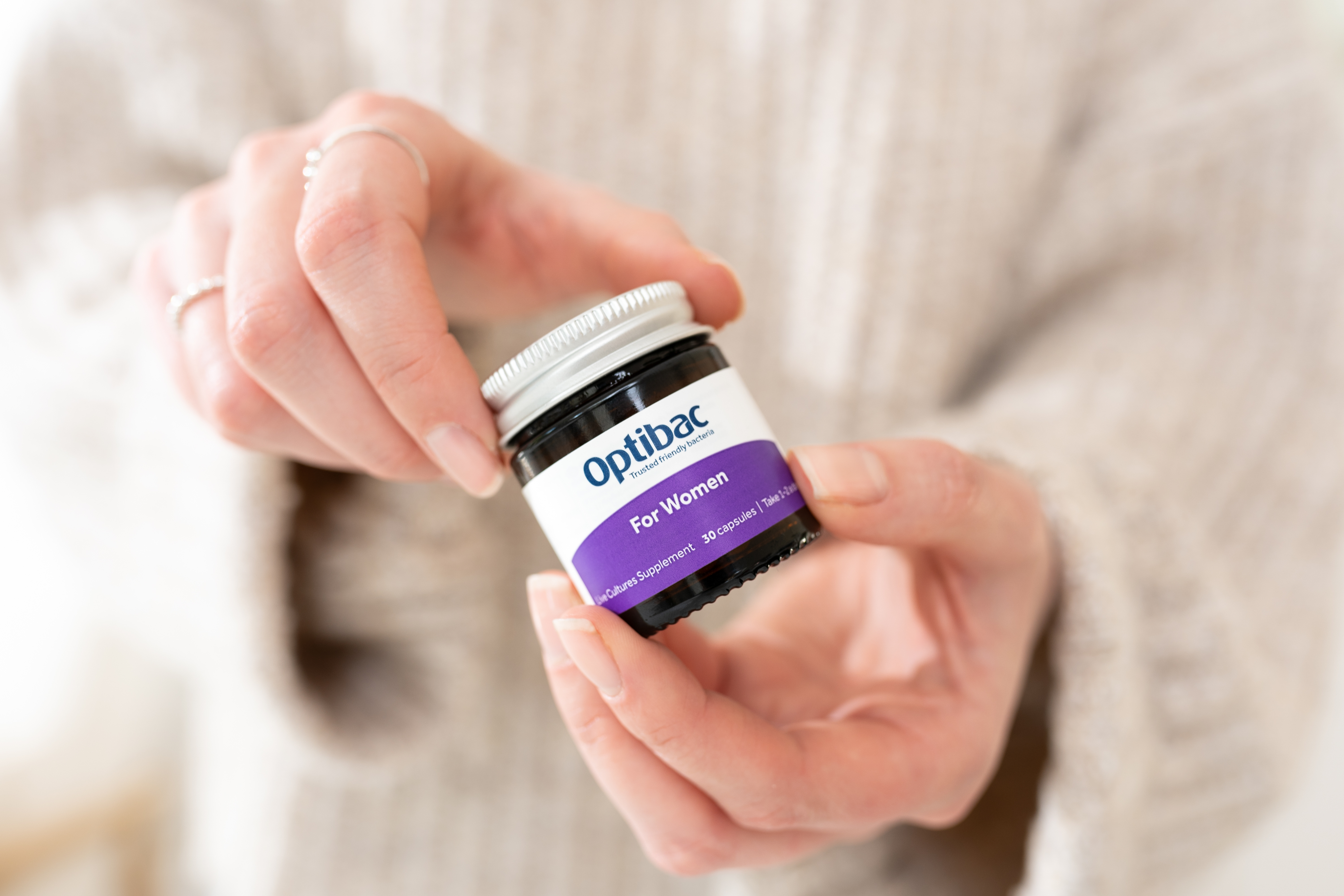 A person holding a small jar labeled "Optibac For Women," a supplement with 30 capsules, against a blurred background.