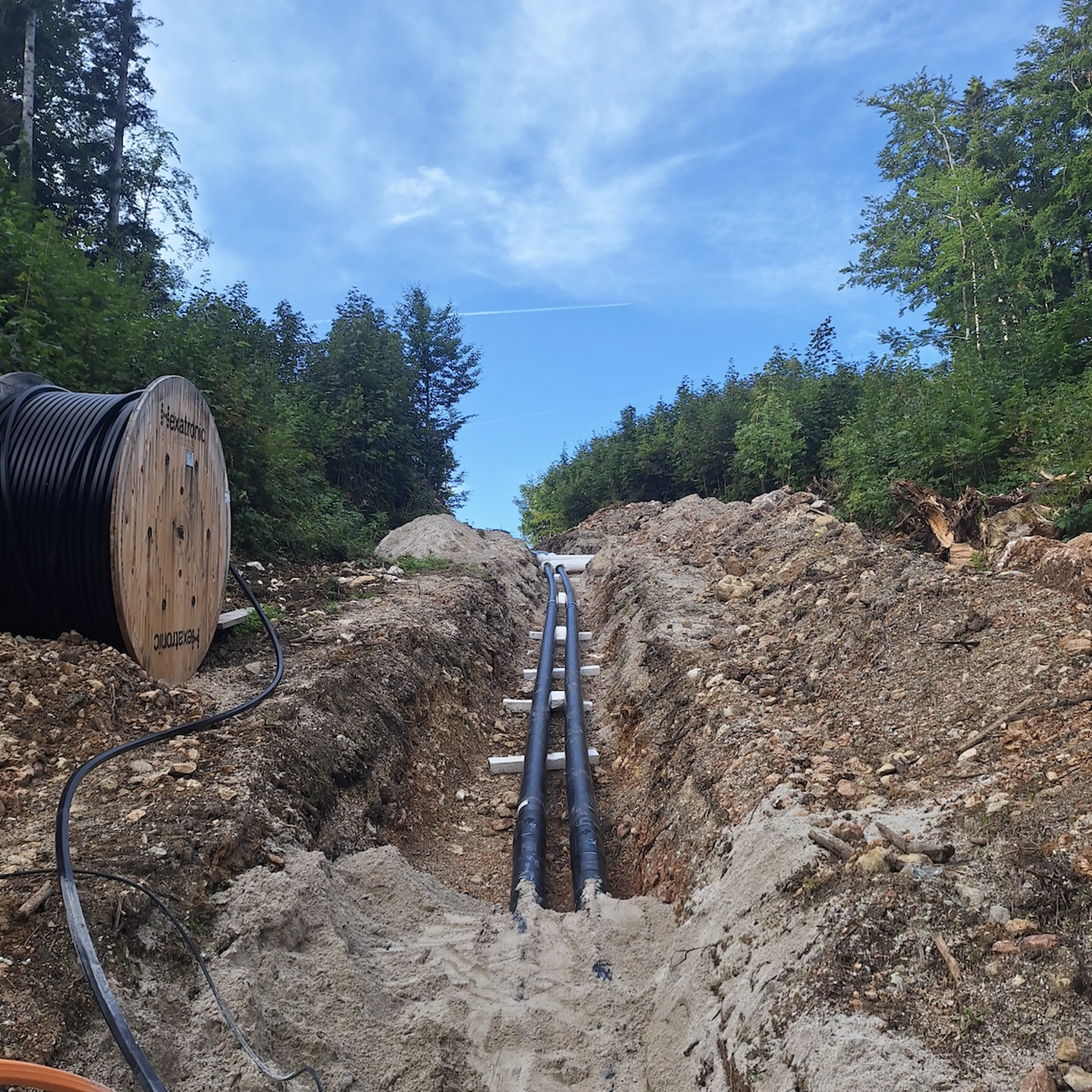Large cables being installed in a forest trench, with a cable spool nearby, beneath a clear blue sky.