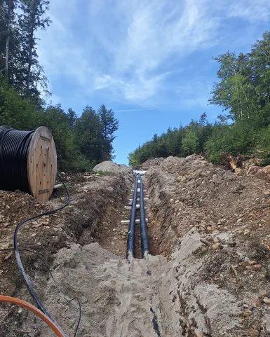 Large cables being installed in a forest trench, with a cable spool nearby, beneath a clear blue sky.