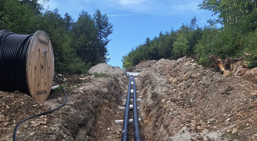 Large cables being installed in a forest trench, with a cable spool nearby, beneath a clear blue sky.