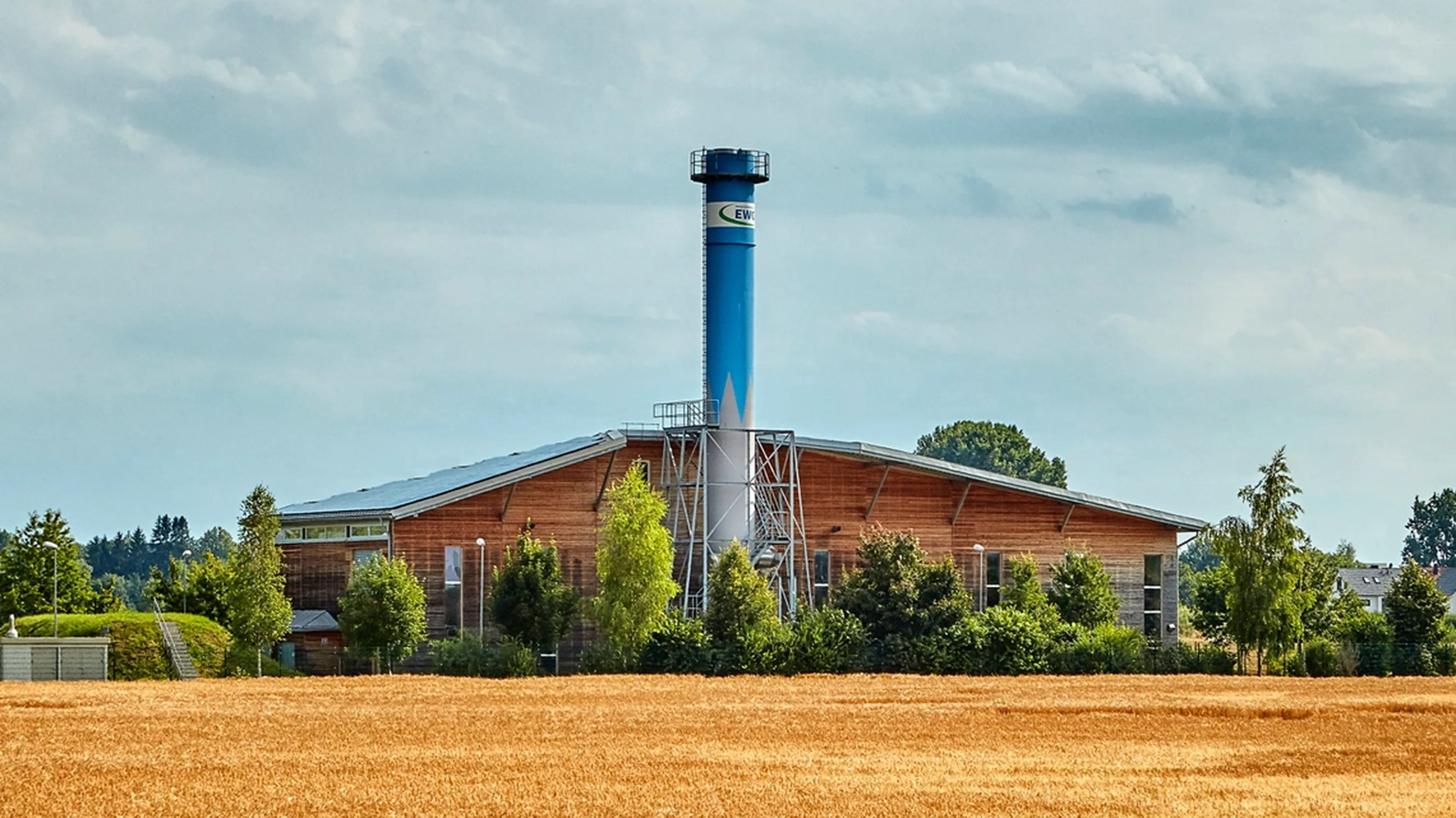 A modern, large industrial building with a tall, blue chimney, surrounded by trees, set against a cloudy sky and a golden field in the foreground.