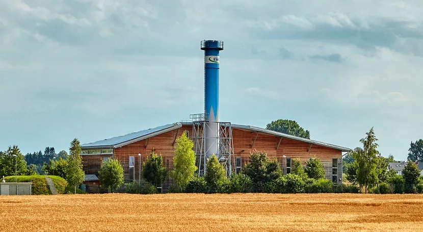 A modern, large industrial building with a tall, blue chimney, surrounded by trees, set against a cloudy sky and a golden field in the foreground.