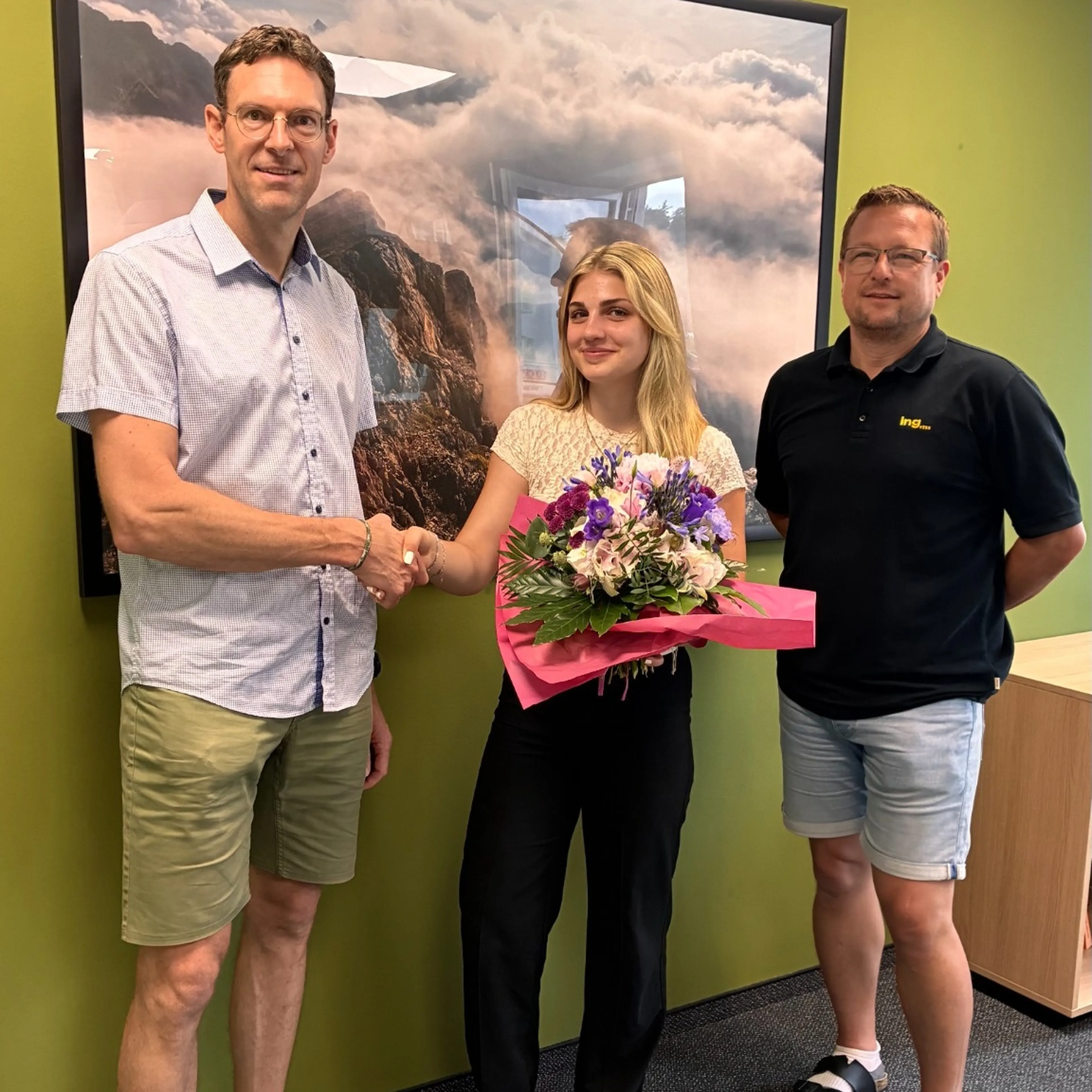 Three people standing in an office; a woman holds a bouquet and is shaking hands with a man. Another man stands beside them, smiling.