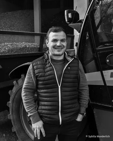 Smiling man in a puffer vest stands beside a vehicle, with stacked materials in the background. Black and white photo.