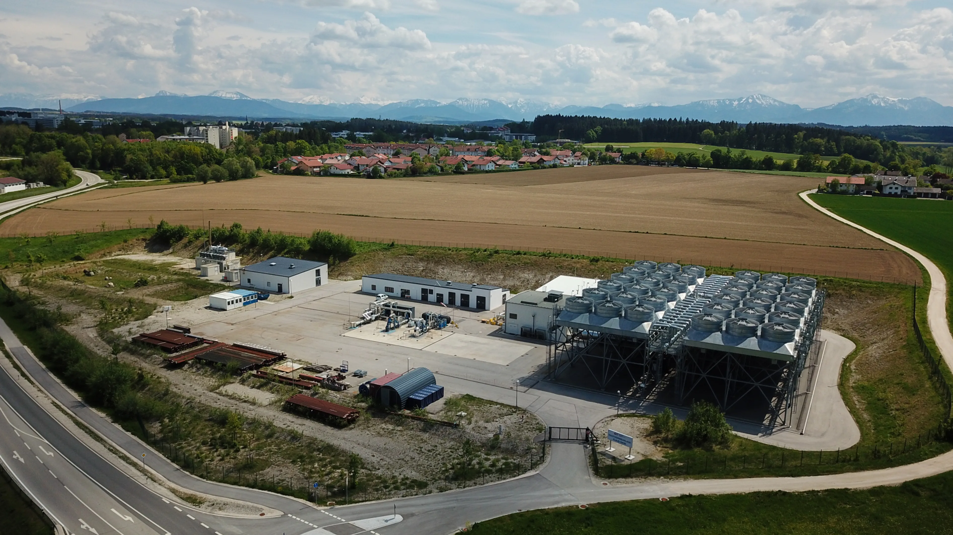 Aerial view of an industrial facility with large cooling units, surrounded by fields and roads, with mountains visible in the distance.