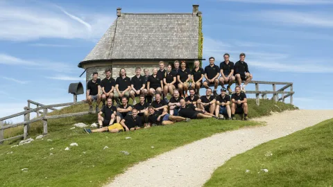 A group of people in black shirts sit and stand on a grassy hill by a stone building, with a blue sky in the background.