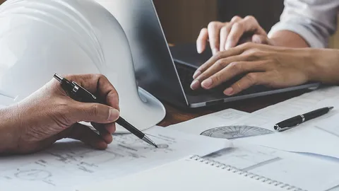 Two people working at a desk with architectural plans, a laptop, a hard hat, and a protractor, one pointing with a pen.