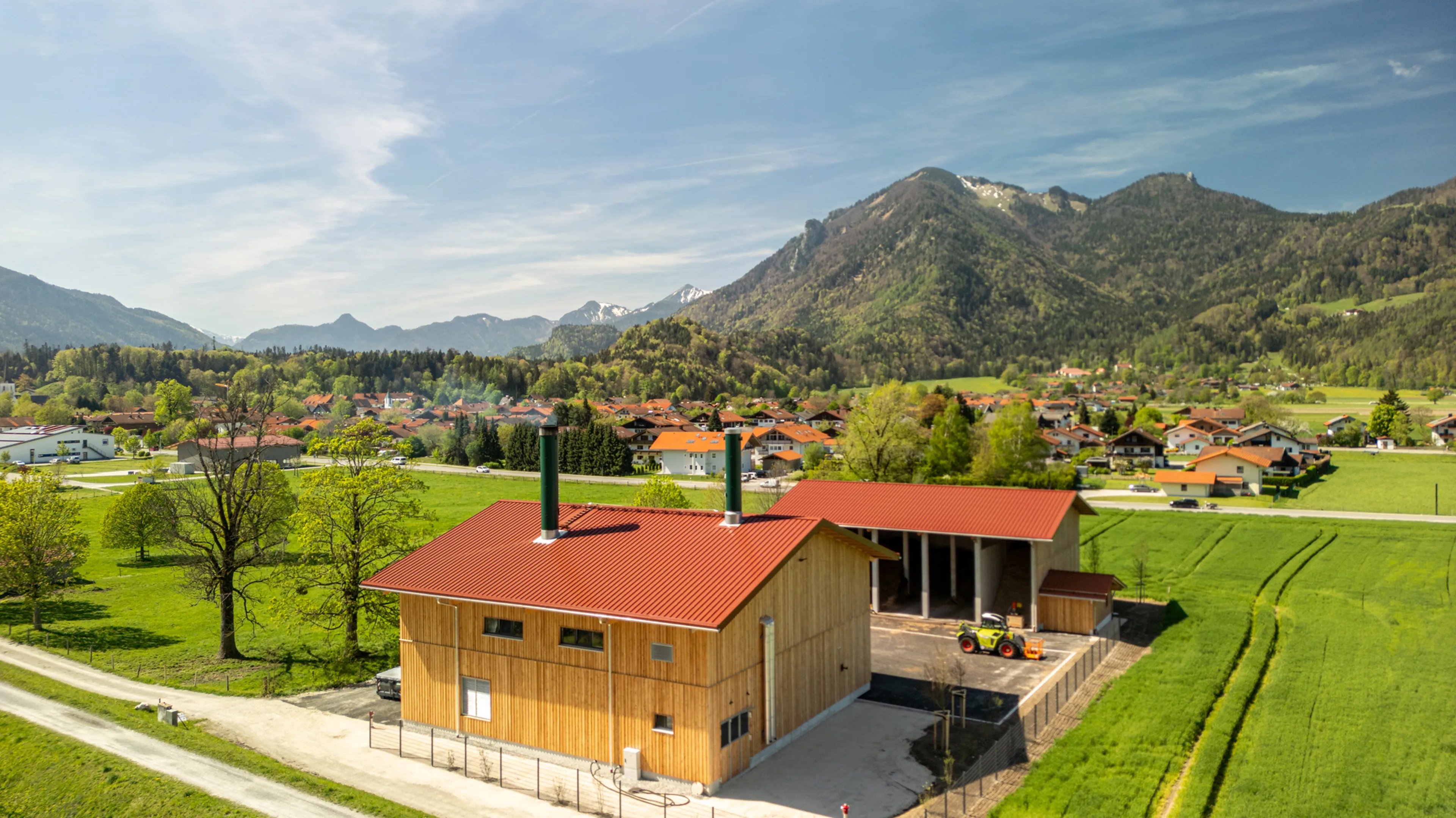 Aerial view of a rural landscape with a red-roofed wooden building, green fields, scattered houses, and mountains in the background under a blue sky.