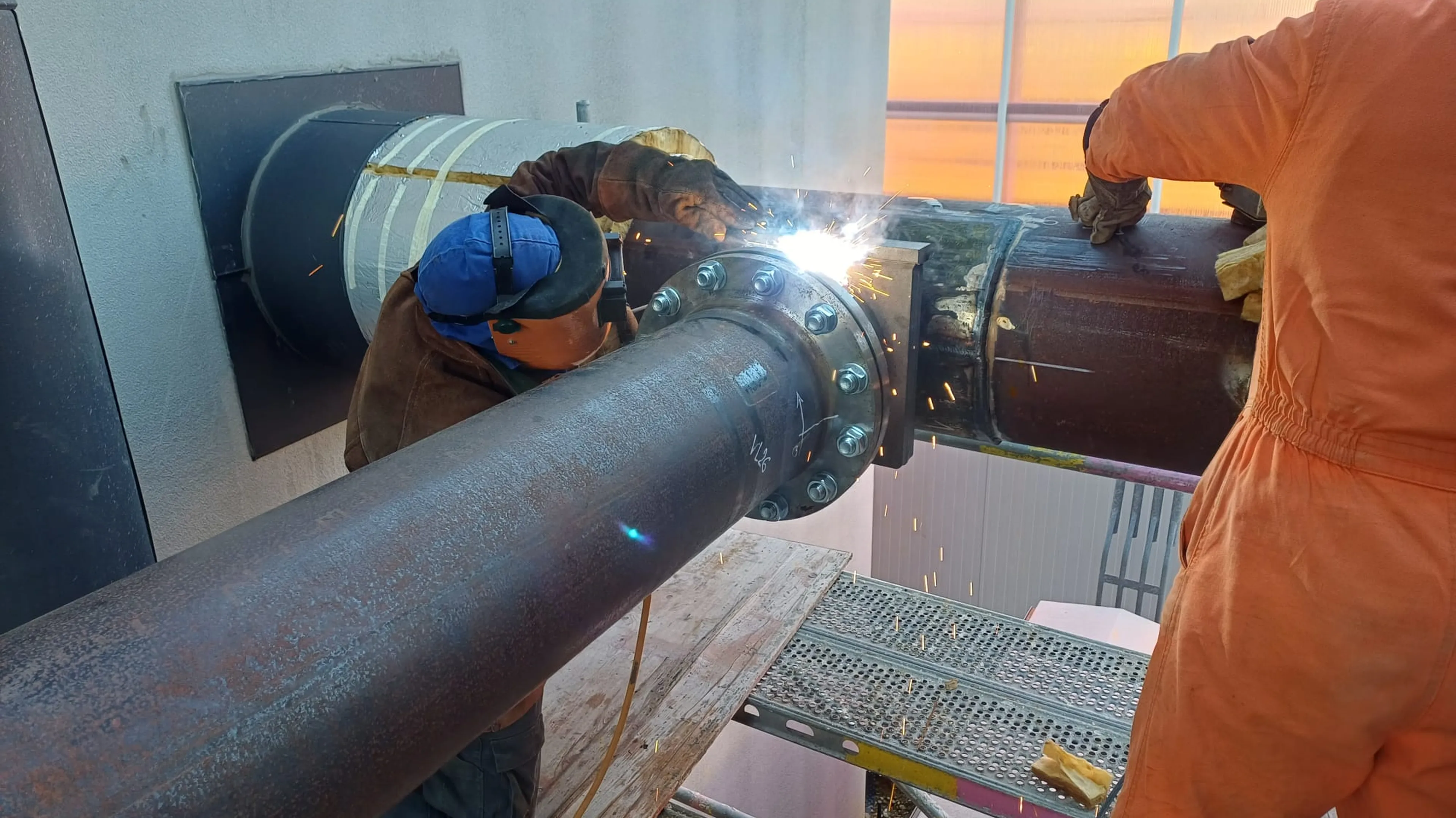 Two workers weld large pipes on scaffolding, with sparks flying and a sunset in the background.