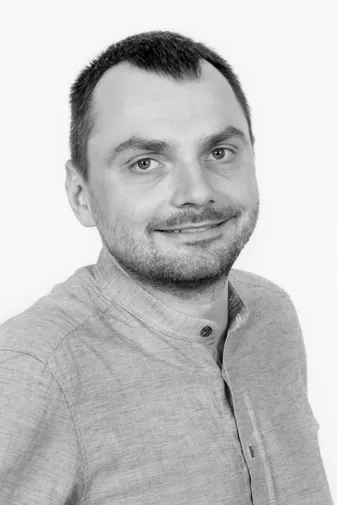 Smiling man with short dark hair and a beard, wearing a light-colored button-up shirt, poses against a plain white background.