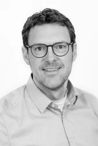 A man with short hair, wearing glasses and a button-up shirt, smiles in a black and white portrait.