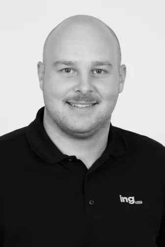 Smiling man with a buzz cut wearing a black polo shirt with "ing kess" logo, against a plain background.