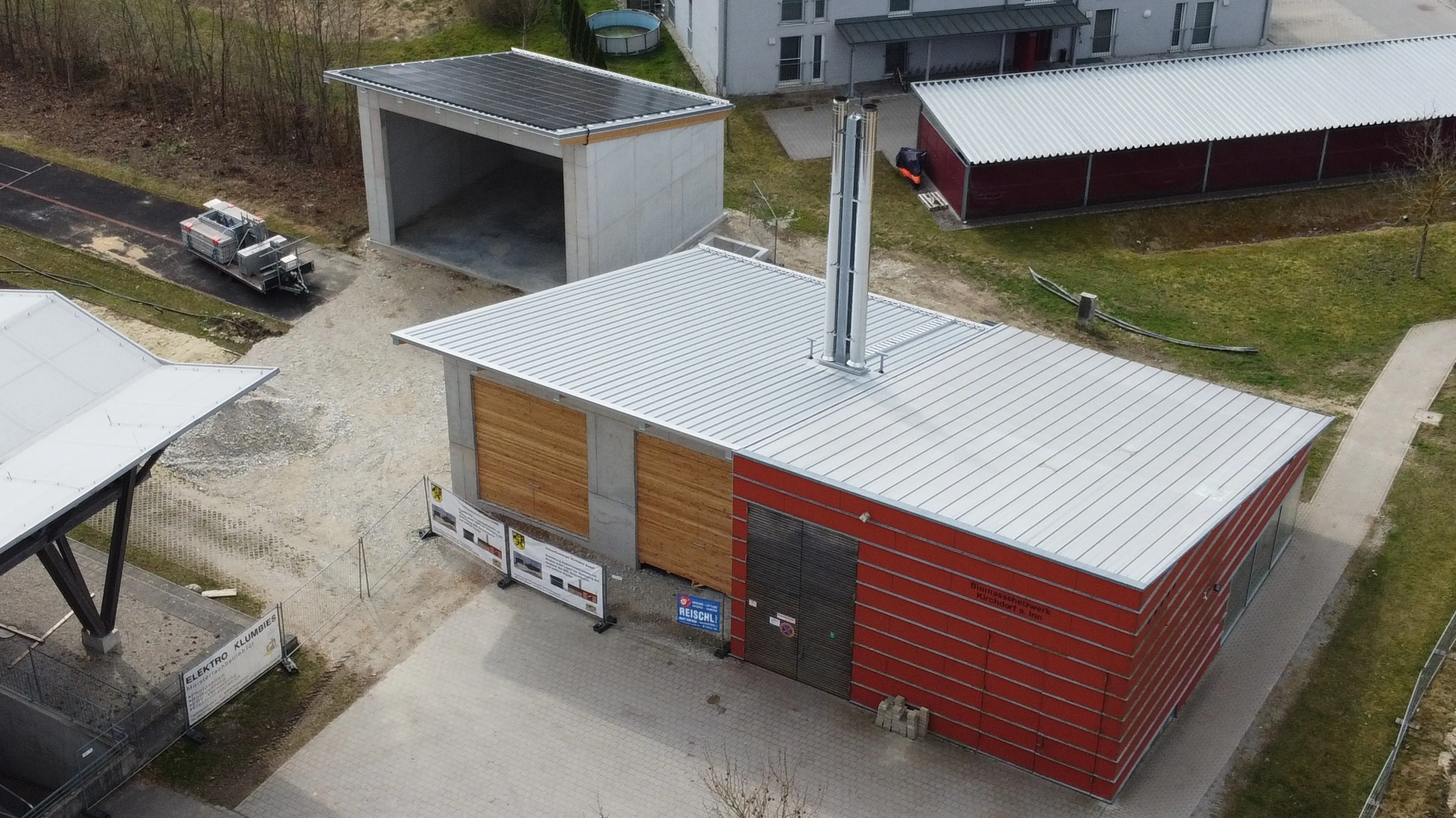 Aerial view of a modern building with red facade and metal roof, surrounded by a paved area, pathway, and parked vehicle.