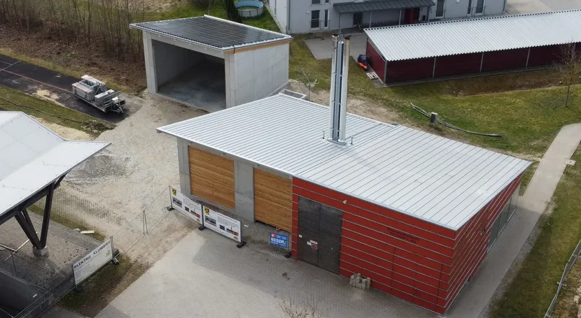 Aerial view of a modern building with red facade and metal roof, surrounded by a paved area, pathway, and parked vehicle.