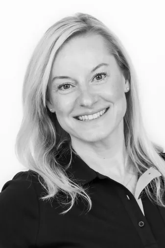 Black and white portrait of a smiling person with long, light hair, wearing a dark collared shirt, looking directly at the camera.