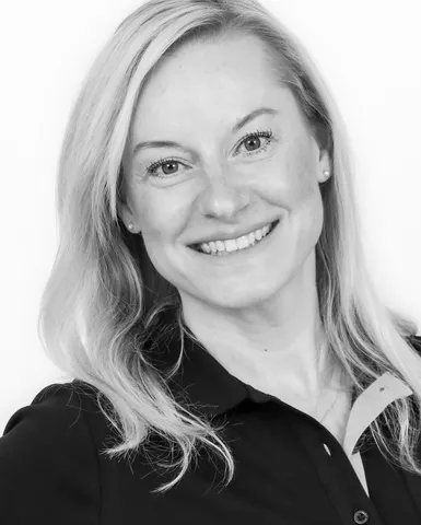 Black and white portrait of a smiling person with long, light hair, wearing a dark collared shirt, looking directly at the camera.