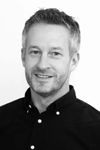 Man with short gray hair and a beard, wearing a dark button-up shirt, smiles at the camera against a plain white background.